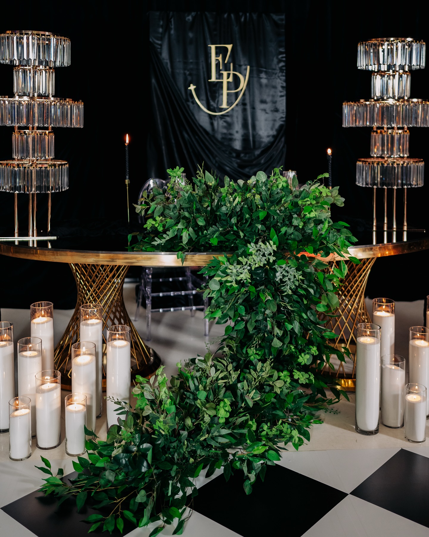 Gold sweetheart table draped in cascading greenery, flanked by crystal tiered stands and white pillar candles, before a black drape with a gold monogram on a black-and-white checkered floor.