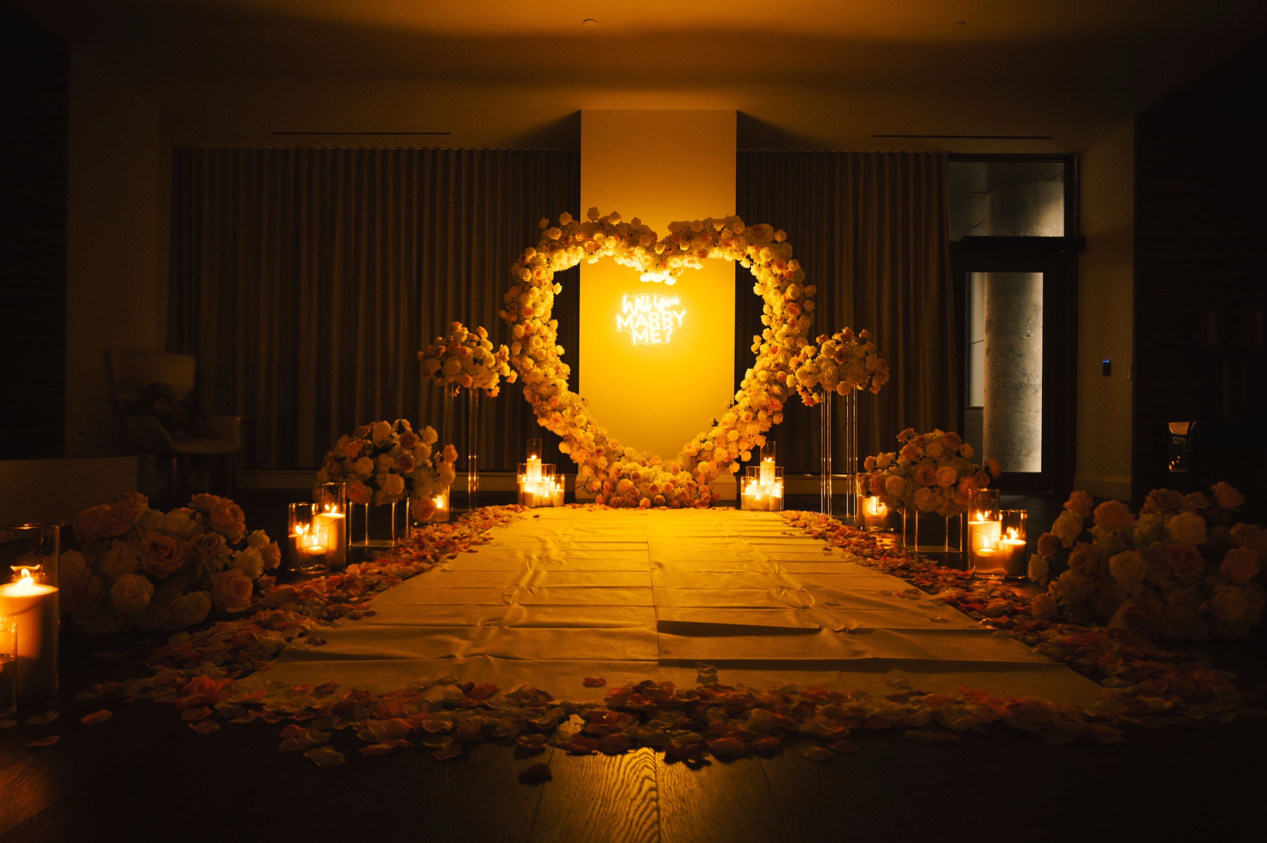Candlelit floral heart arch with a neon “Will you marry me?” sign at the end of a long aisle of rose petals, flanked by tall arrangements and glass candle holders.