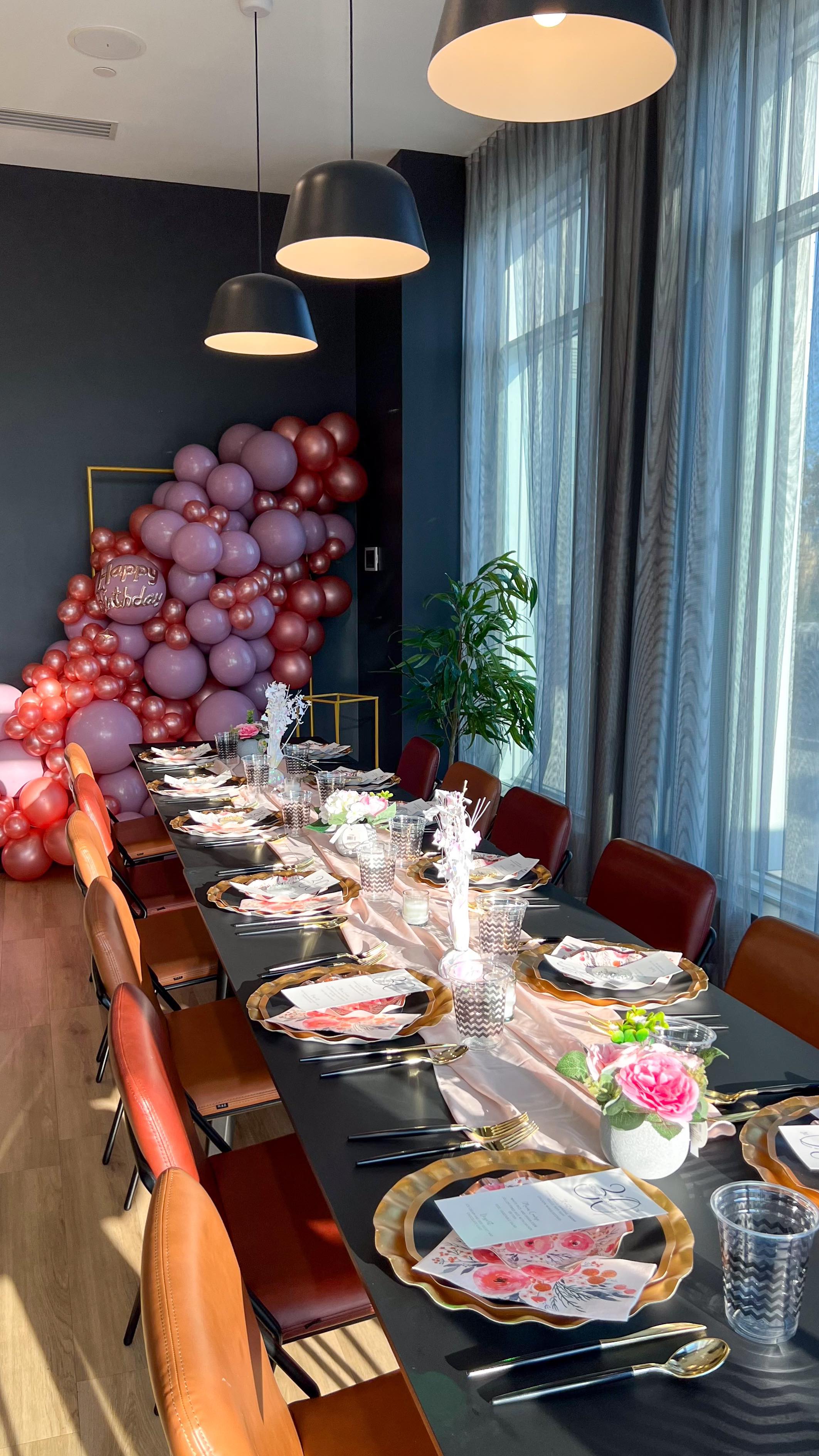 Long dinner table set with gold chargers, pink menus, and pink roses, leading to a pink and burgundy balloon garland beside tall windows.