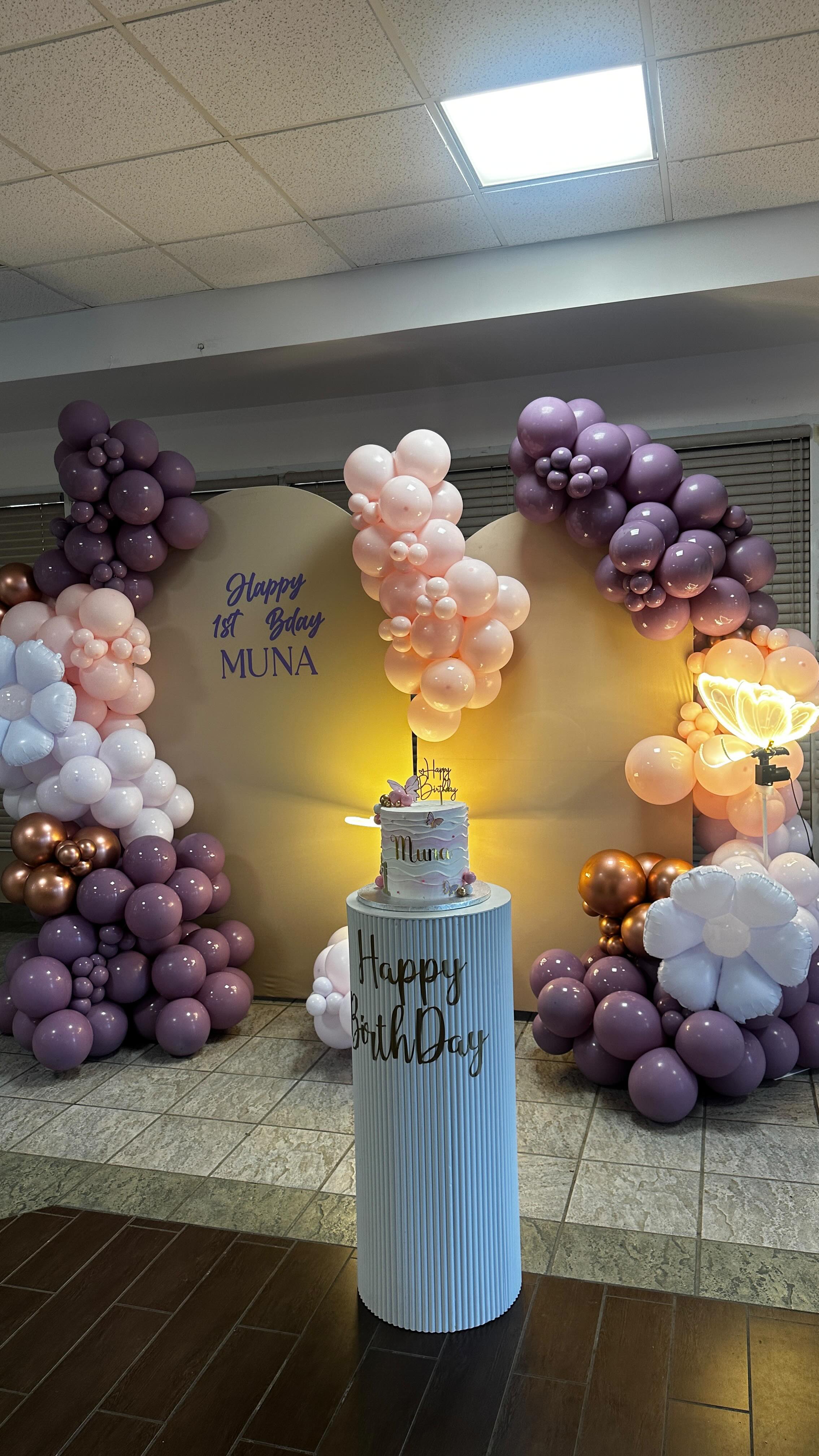Mauve, blush, and copper balloon garlands framing an arched backdrop, with a celebration cake on a fluted white pedestal.